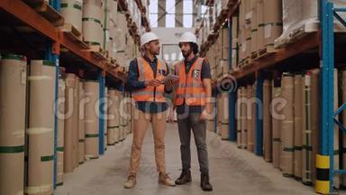 两名年轻男工人站在配送仓库，身穿白色硬帽和反光夹克，检查库存